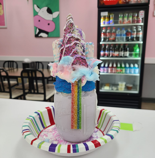A colorful milkshake topped with whipped cream, cotton candy, and a rainbow, served in a mason jar on a paper plate.