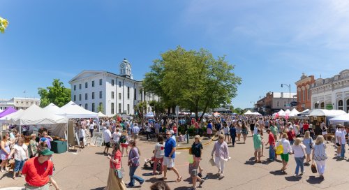 A bustling outdoor market scene with crowds, tents, and a historic building under a clear blue sky.
