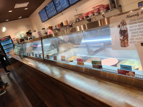 A brightly lit ice cream shop with a glass display case showcasing various flavors and toppings. Customers are in line.