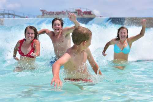 Four children play in a wave pool, splashing water and smiling, with a water park in the background.