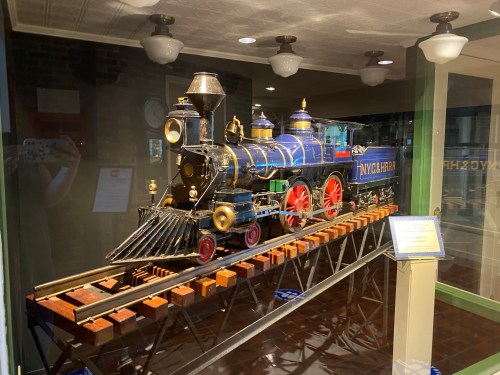 A vintage model steam locomotive displayed in a glass case, featuring blue and gold detailing with red wheels.