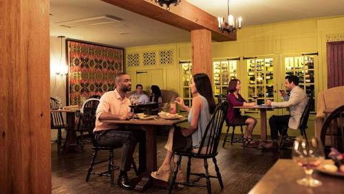 A cozy restaurant scene with couples dining at wooden tables, warm lighting, and a wine display in the background.