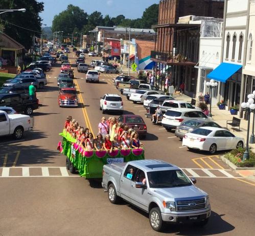 A parade float with people in colorful costumes travels down a busy street lined with cars and shops.