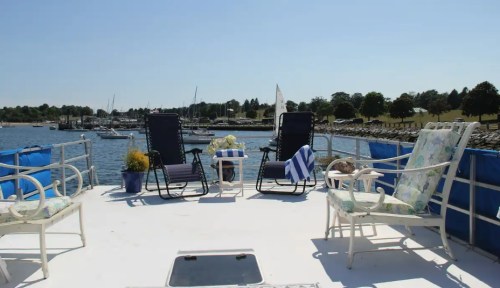 Two chairs and a small table on a boat deck, overlooking a marina with sailboats and trees in the background.