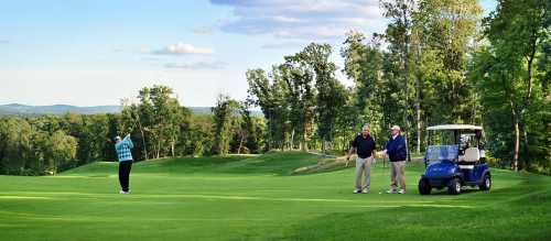 A golfer swings on a lush green course while two men watch from a golf cart nearby, surrounded by trees and hills.