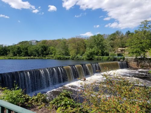 A serene waterfall cascades over a dam, surrounded by lush greenery and a clear blue sky with scattered clouds.