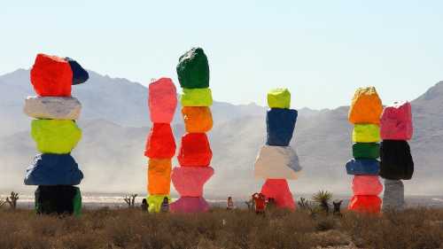 Colorful stacked boulders in a desert landscape, with people nearby and mountains in the background.