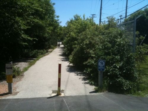 A tree-lined path leads into the distance, with a barrier and signs on either side, under a clear blue sky.