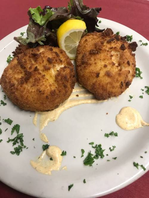 Two golden-brown crab cakes on a plate, garnished with lettuce, a lemon wedge, and drizzled with sauce.