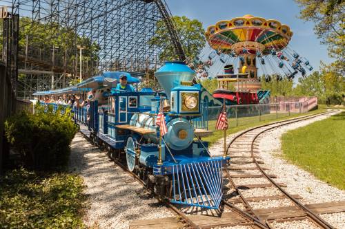 A blue steam train with flags travels along a track near a colorful amusement park ride in a sunny setting.