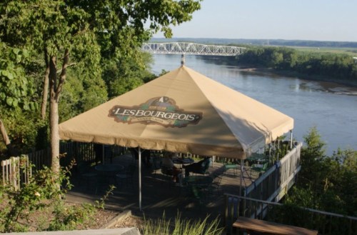 A riverside patio with a large tan canopy, overlooking a river and bridge, surrounded by trees.
