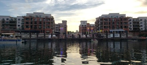 A waterfront view of modern buildings with balconies, reflecting in the water under a colorful sunset sky.