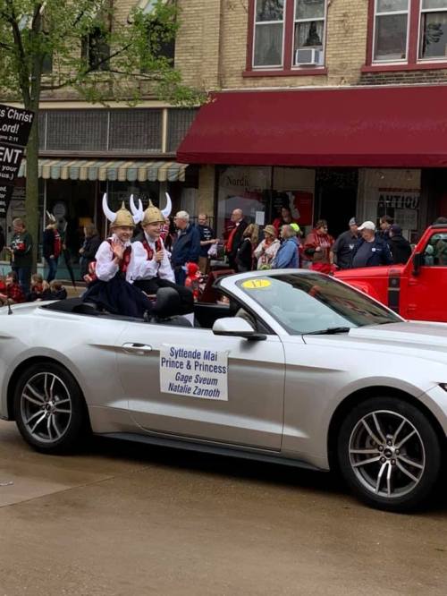 A convertible car in a parade with two people dressed in traditional attire, waving to the crowd.