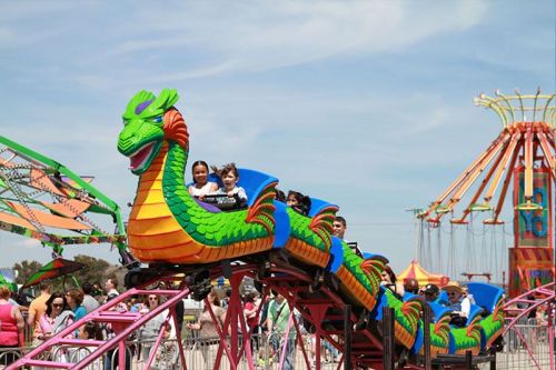 Children enjoy a colorful dragon-themed roller coaster at a fair, with vibrant rides in the background.