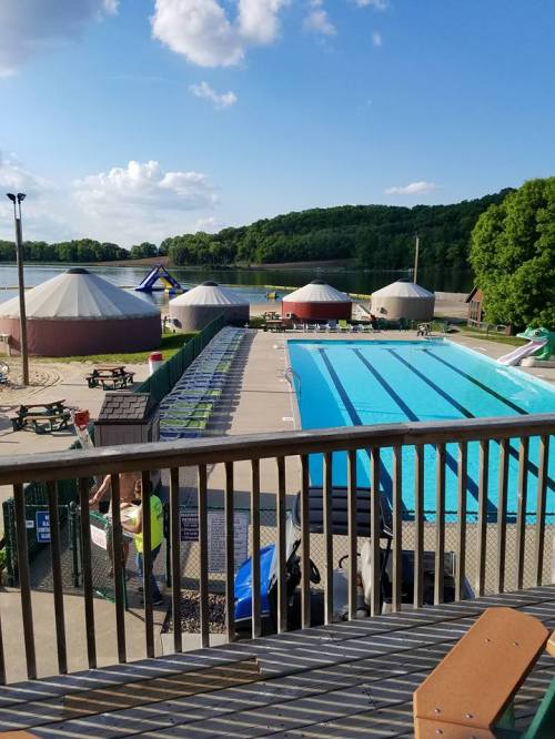 View of a swimming pool surrounded by lounge chairs, with tents and a lake in the background under a sunny sky.