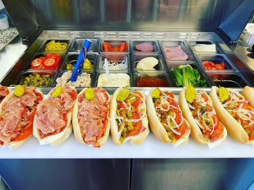 A row of six sub sandwiches filled with meats, cheese, and toppings, displayed in front of a condiment station.