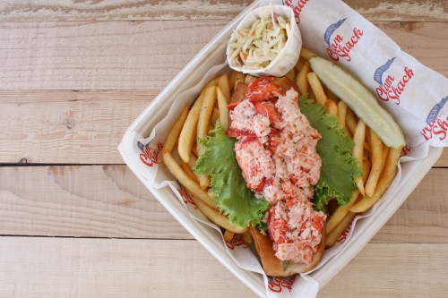 A lobster roll topped with lobster meat on a bed of lettuce, served with fries, coleslaw, and a pickle.