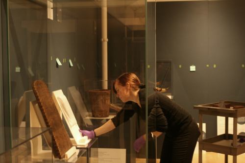 A museum worker in gloves examines an exhibit behind glass in a dimly lit gallery.