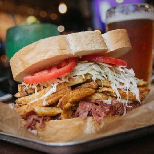A towering sandwich with layers of fried potatoes, shredded lettuce, and tomato, served with a drink in the background.