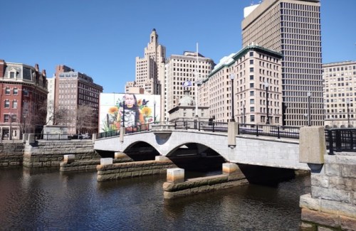 A stone bridge over a river with a mural and city buildings in the background under a clear blue sky.