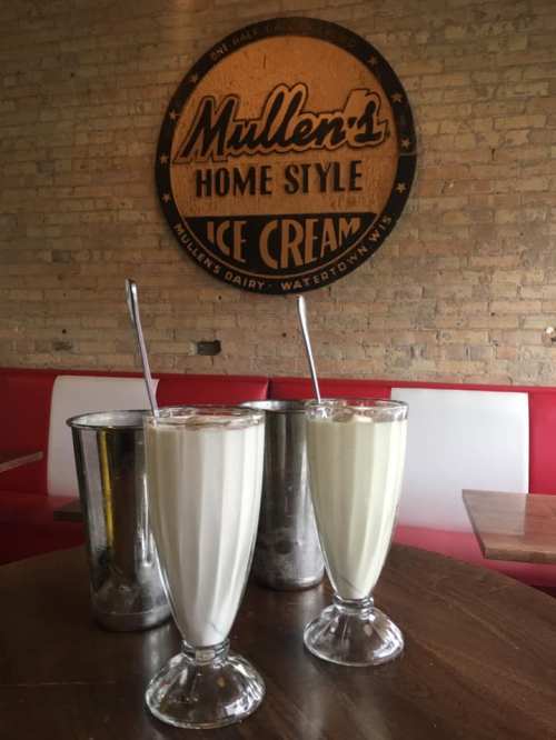 Two tall glasses of milkshakes sit on a table, with metal containers in the background and a sign for Mullen's Ice Cream.