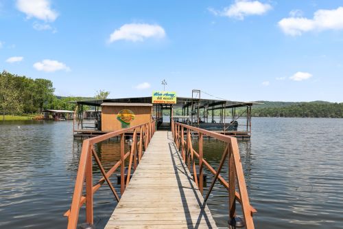 A wooden dock leads to a lakeside building surrounded by water and greenery under a clear blue sky.