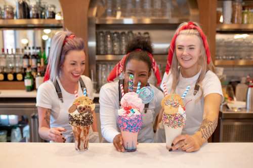 Three smiling women in aprons hold colorful milkshakes topped with sweets, standing behind a bar with drinks in the background.