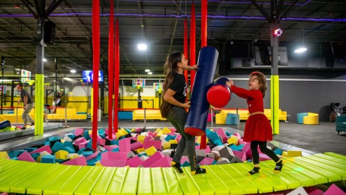 Two children play on a colorful trampoline area, one holding a foam pole while the other prepares to hit a foam ball.