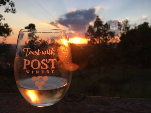 A glass of wine with "Toast with Post Winery" etched on it, set against a vibrant sunset and trees in the background.