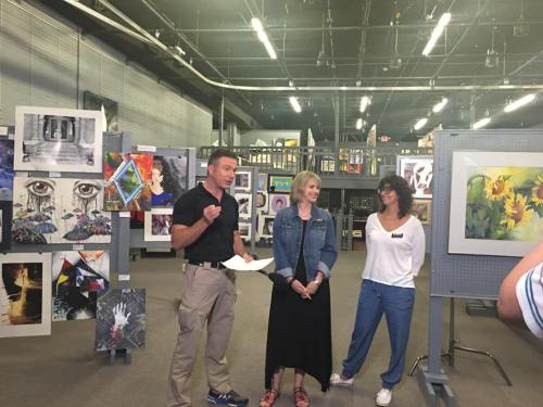 A man speaks while two women listen in an art gallery filled with various artworks on display.