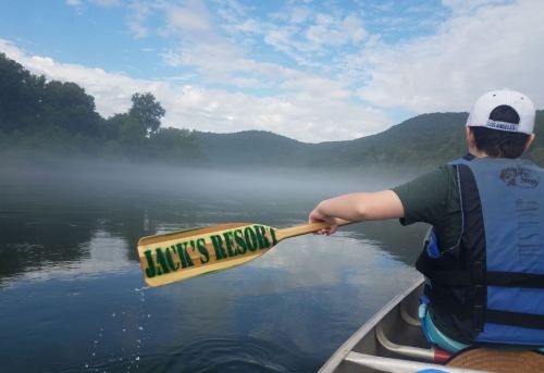 A person in a canoe holds a paddle with "JACK'S RESORT" written on it, surrounded by misty water and mountains.