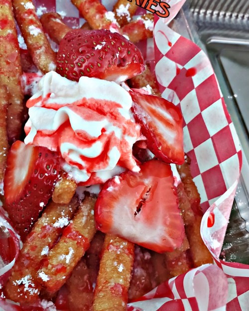 A close-up of a dessert featuring fried dough sticks topped with strawberries, whipped cream, and strawberry sauce.