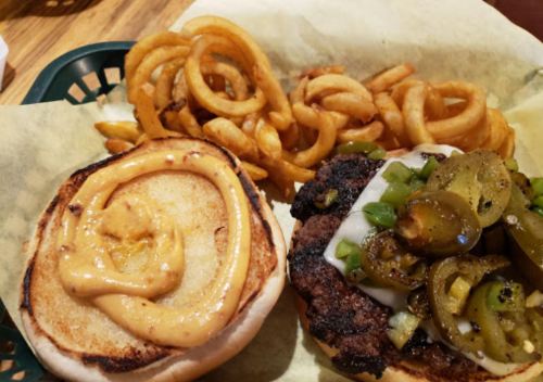A close-up of a burger with jalapeños and sauce on a toasted bun, served with curly fries on the side.