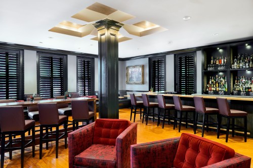 A modern bar area with dark wood accents, high stools, and red upholstered chairs, featuring a well-stocked liquor shelf.