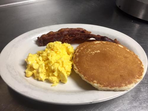 A plate with scrambled eggs, a pancake, and crispy bacon on a stainless steel surface.