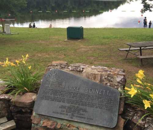 A stone plaque at Lake Bennett, with a calm lake and people in the background, surrounded by green grass and flowers.