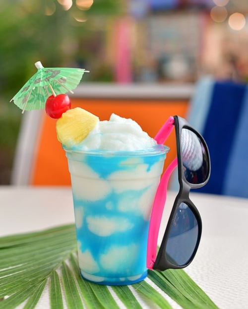 A colorful tropical drink with a cherry, pineapple, and a small umbrella, accompanied by sunglasses on the side.