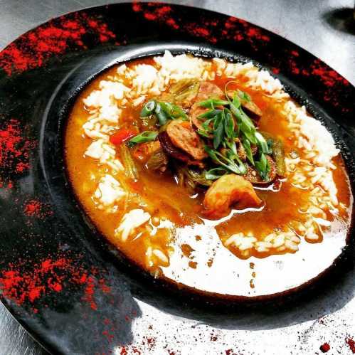 A bowl of flavorful stew with rice, garnished with green onions and a sprinkle of red seasoning on a black plate.