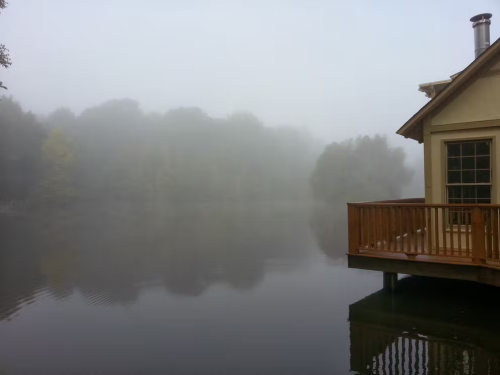 A foggy scene by a calm lake, with a wooden deck and a house partially visible in the mist.