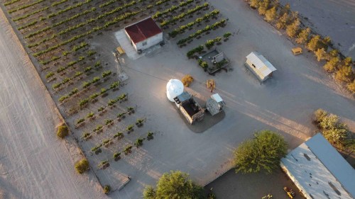 Aerial view of a rural area with vineyards, buildings, and a dome structure surrounded by dirt paths and trees.