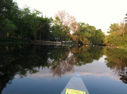 A calm lake reflecting trees and a shoreline, viewed from the front of a canoe.