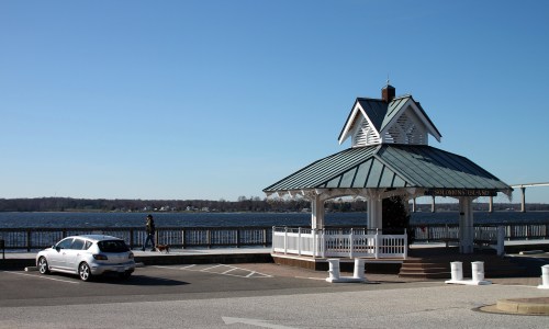 A waterfront gazebo with a car parked nearby, overlooking a calm river on a clear day.
