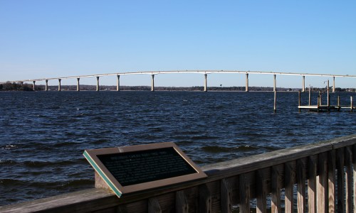 A view of a bridge spanning a river, with a wooden railing and informational plaque in the foreground.