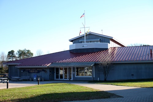 A modern building with a red roof, featuring flags and a grassy area in front, under a clear blue sky.