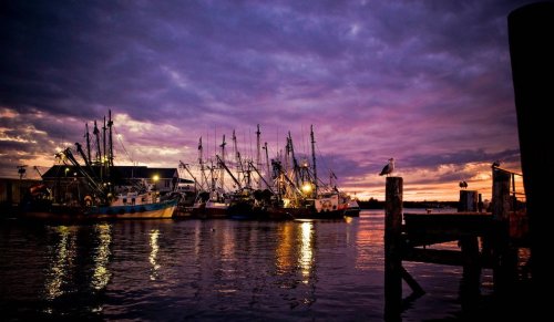 Fishing boats docked at sunset, reflecting in calm waters under a colorful sky with clouds.