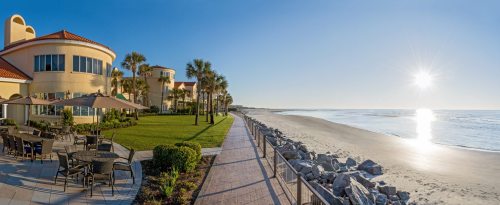 A sunny beachside view with palm trees, a walkway, and a building overlooking the ocean at sunrise.