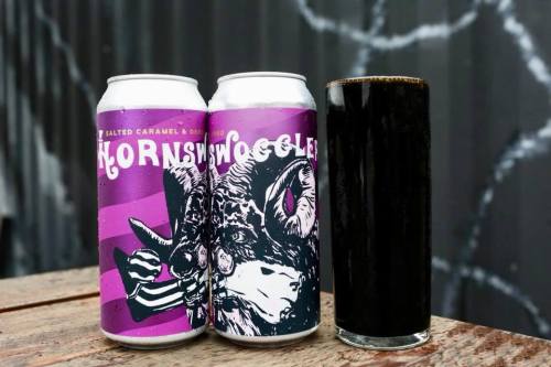 Two cans of "Hornswoggle" beer with a dark glass of beer beside them, set on a wooden table against a textured background.