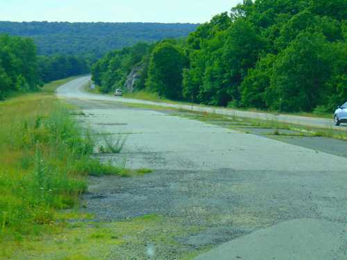 A winding road surrounded by lush greenery, with a patchy asphalt surface and a few cars in the distance.