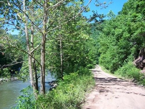 A dirt path lined with trees runs alongside a river, surrounded by lush green hills under a clear blue sky.