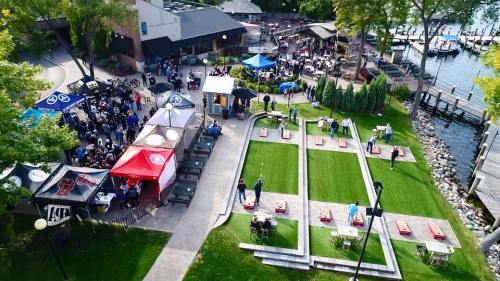 Aerial view of a lively outdoor event with tents, games, and a crowd near a waterfront. Green lawns and boats visible.